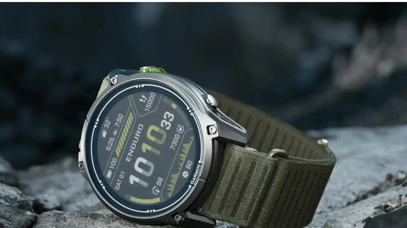 Close-up of a Garmin Enduro 4 titanium sports watch with a dark olive ComfortFit fabric band against a rocky background