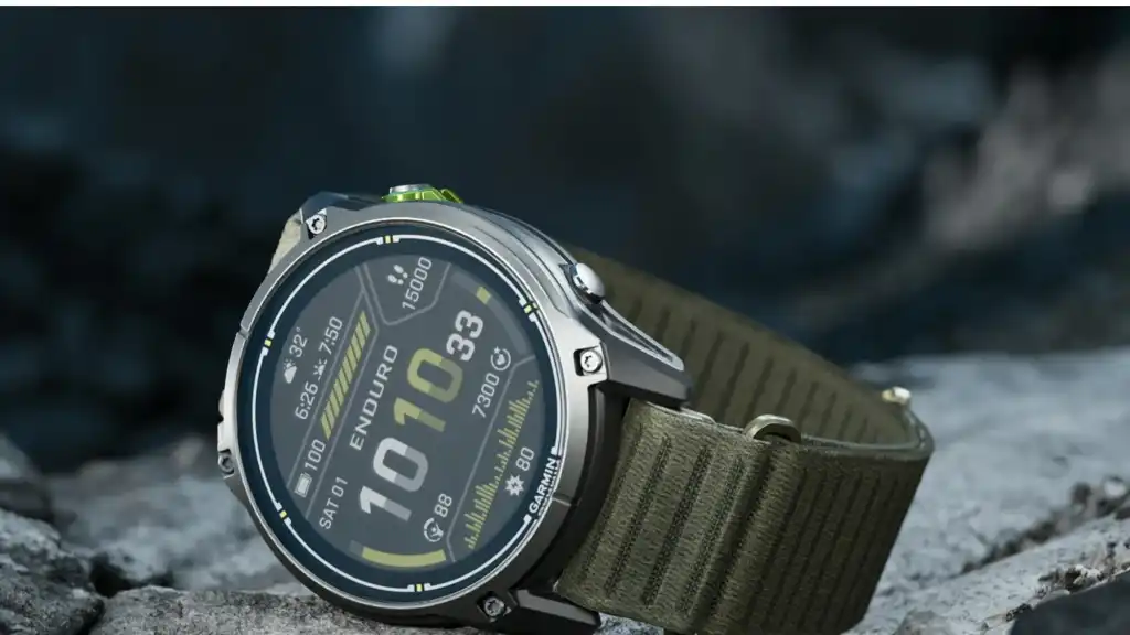 Close-up of a Garmin Enduro 4 titanium sports watch with a dark olive ComfortFit fabric band against a rocky background