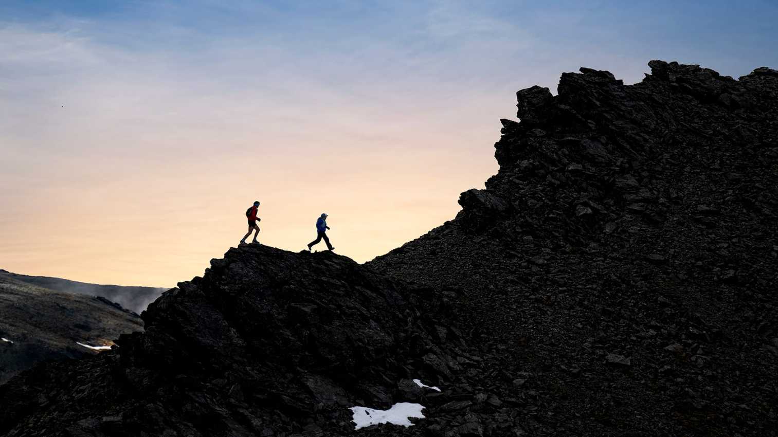 Two trail runners on a rocky mountain ridge against an orange sky