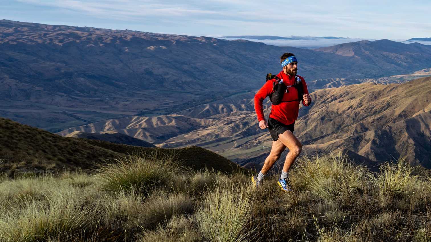 alt="Trail runner wearing a red top and hydration vest on a grassy mountainside with panoramic mountain views