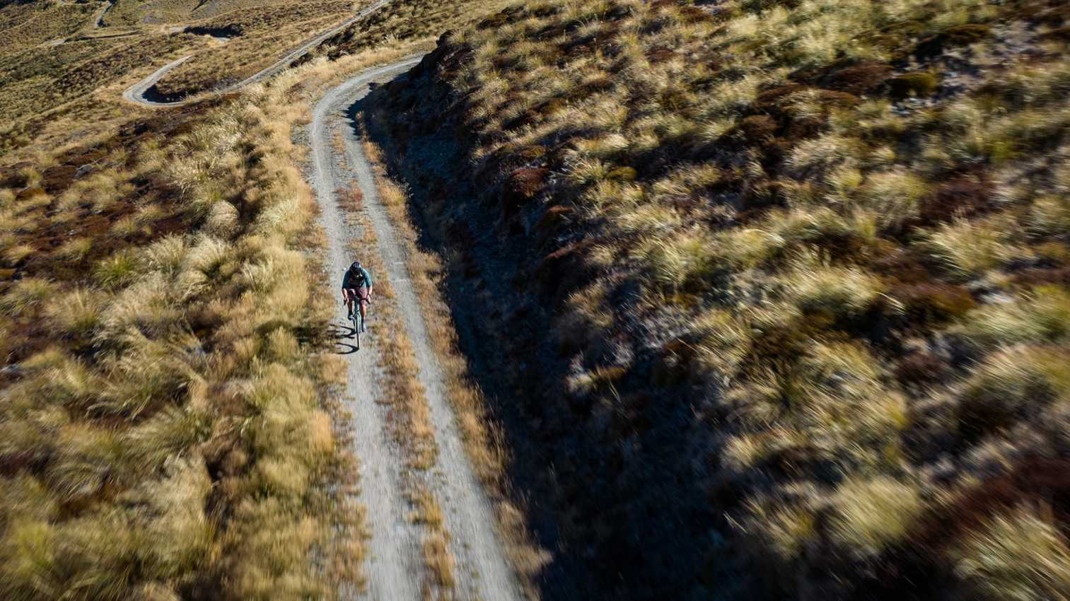 Aerial view of a cyclist on a winding gravel road across a grassy mountainside