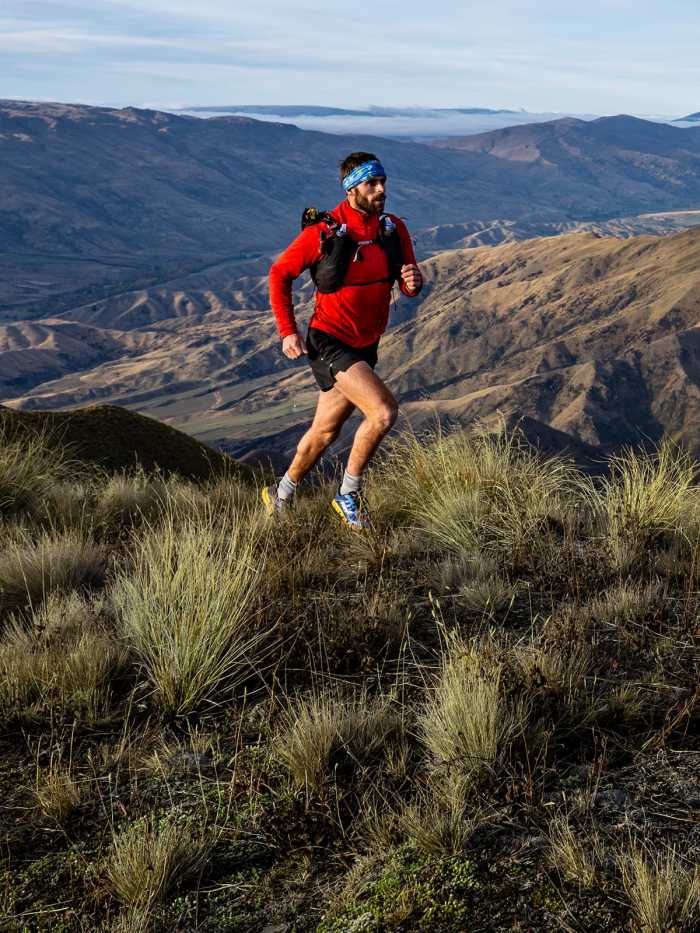 alt="Trail runner wearing a red top and hydration vest on a grassy mountainside with panoramic mountain views