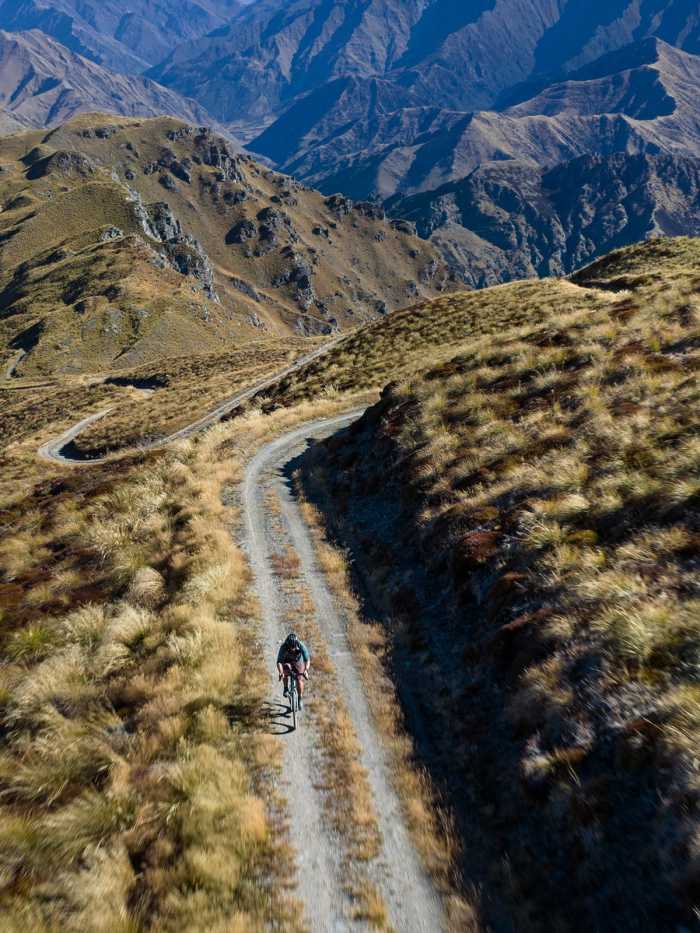 Aerial view of a cyclist on a winding gravel road across a grassy mountainside
