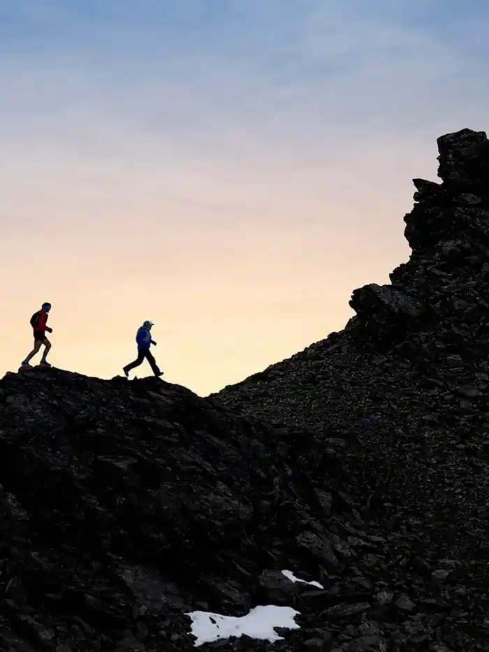 Two trail runners on a rocky mountain ridge against an orange sky